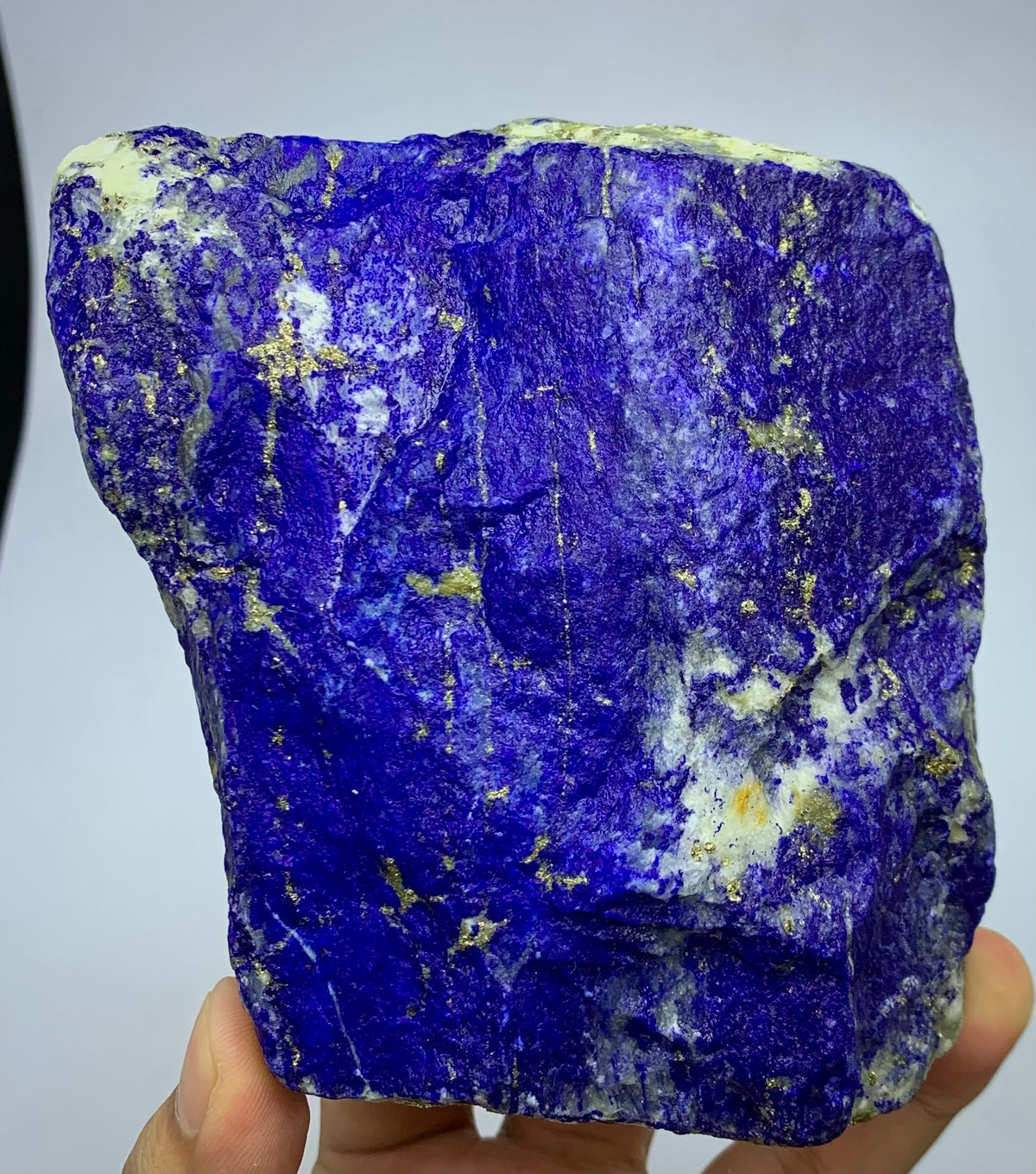 A raw, rough-cut lapis lazuli crystal specimen held in a hand, showcasing its deep blue-violet color with golden pyrite inclusions and white calcite veins.