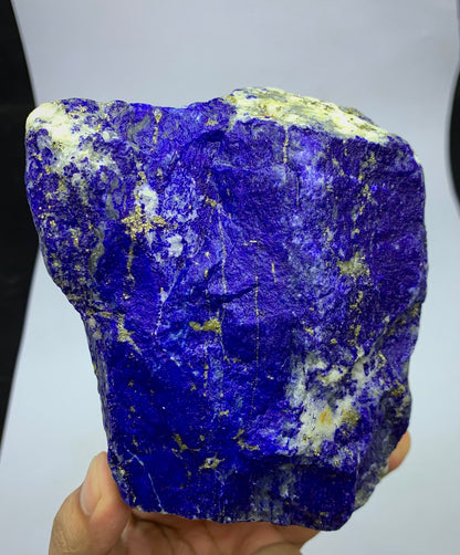 A raw, rough Lapis Lazuli crystal specimen held in a hand, showcasing its deep blue-violet matrix with prominent golden pyrite veins and white calcite inclusions.