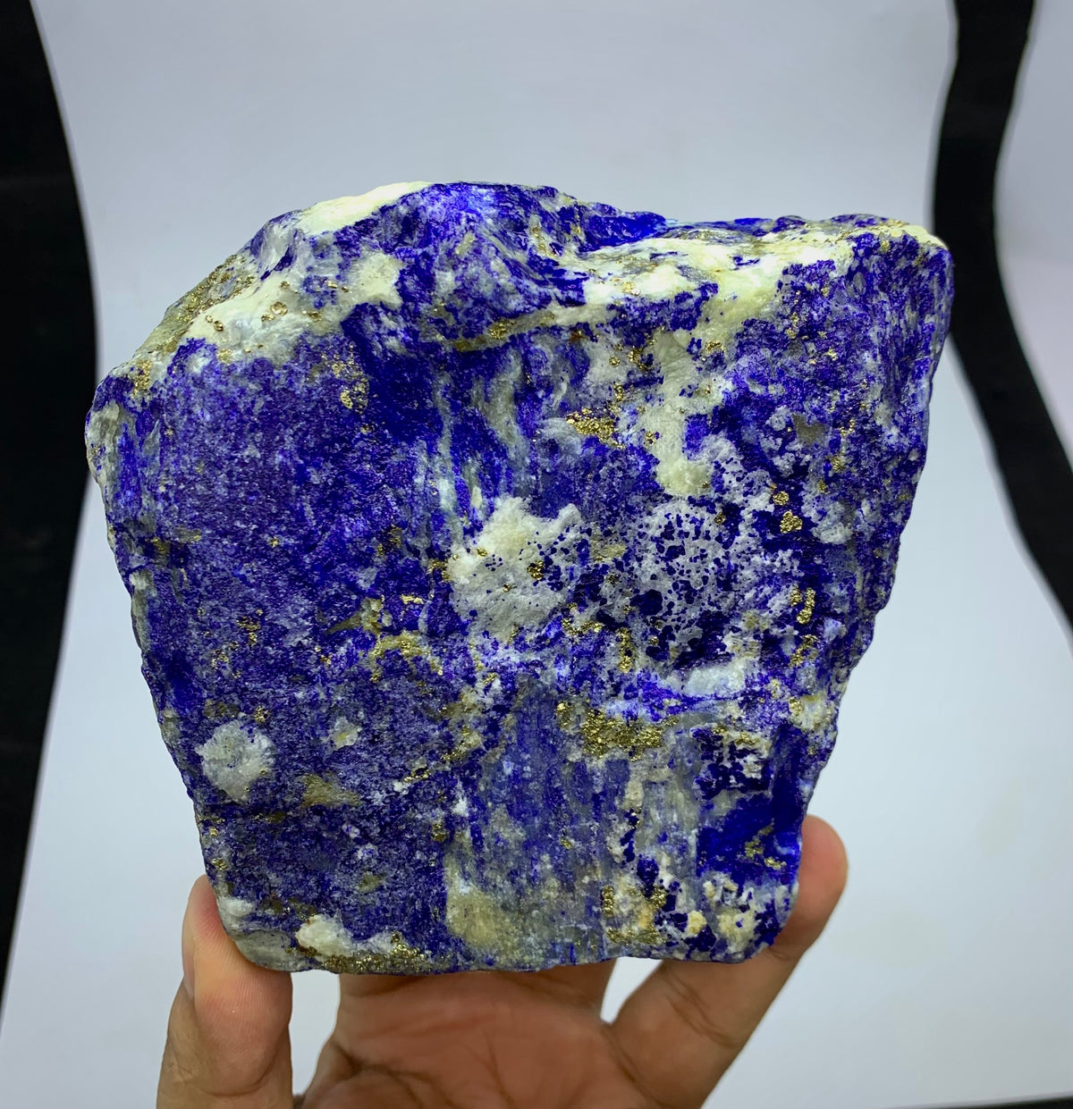 A hand holding a rough, irregularly shaped lapis lazuli crystal with deep blue, white, and gold matrix, set against a white background with black side borders.