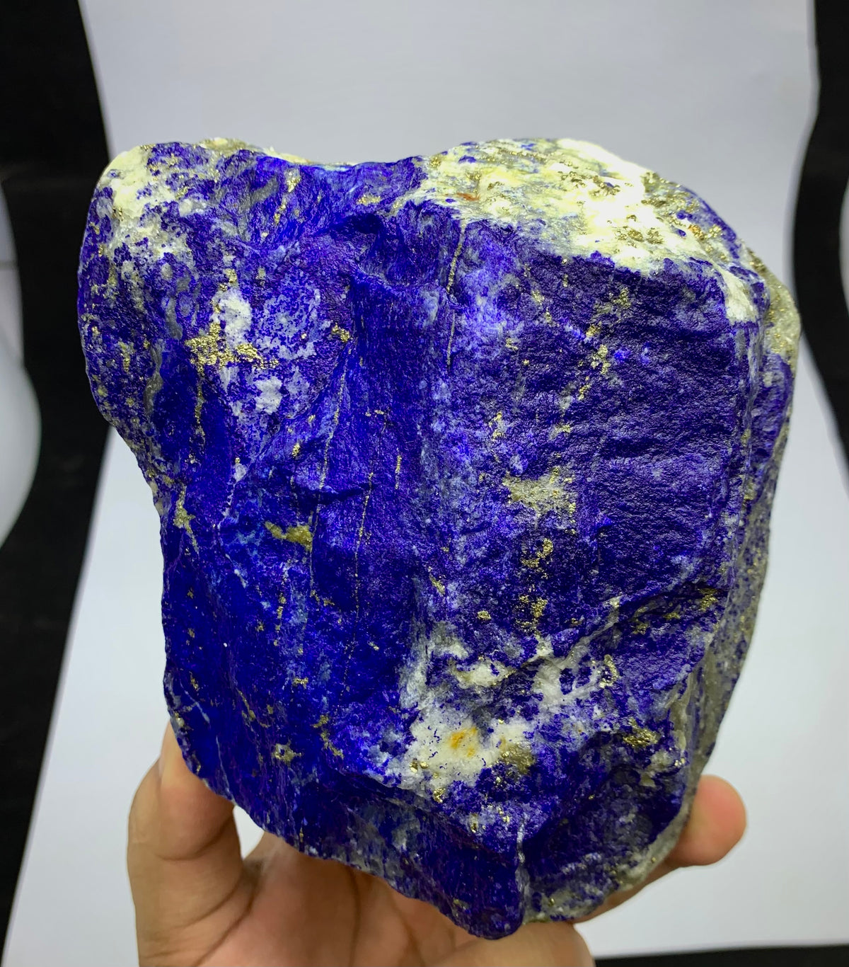 A raw, rough Lapis Lazuli crystal specimen held in a hand, showcasing its deep blue-violet color with natural gold pyrite and white calcite inclusions.