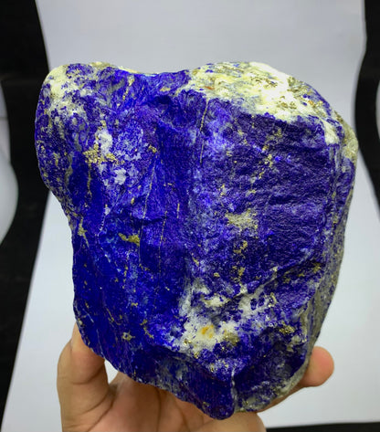 A raw, rough Lapis Lazuli crystal specimen held in a hand, showcasing its deep blue-violet color with natural gold pyrite and white calcite inclusions.