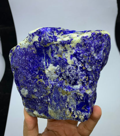 A raw lapis lazuli crystal specimen with deep blue, white, and gold flecks, held in a person's hand against a white and black background.