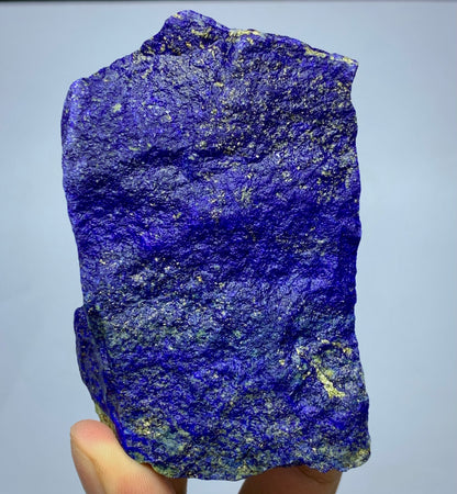 A raw, rough-cut piece of lapis lazuli crystal held in a hand, showcasing its deep blue color with prominent metallic gold pyrite inclusions against a neutral light gray background.