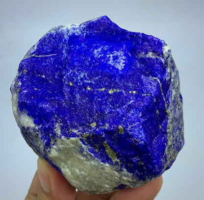 A raw lapis lazuli crystal specimen with deep blue coloration and visible gold pyrite veins, held in a person's hand against a plain light gray background.