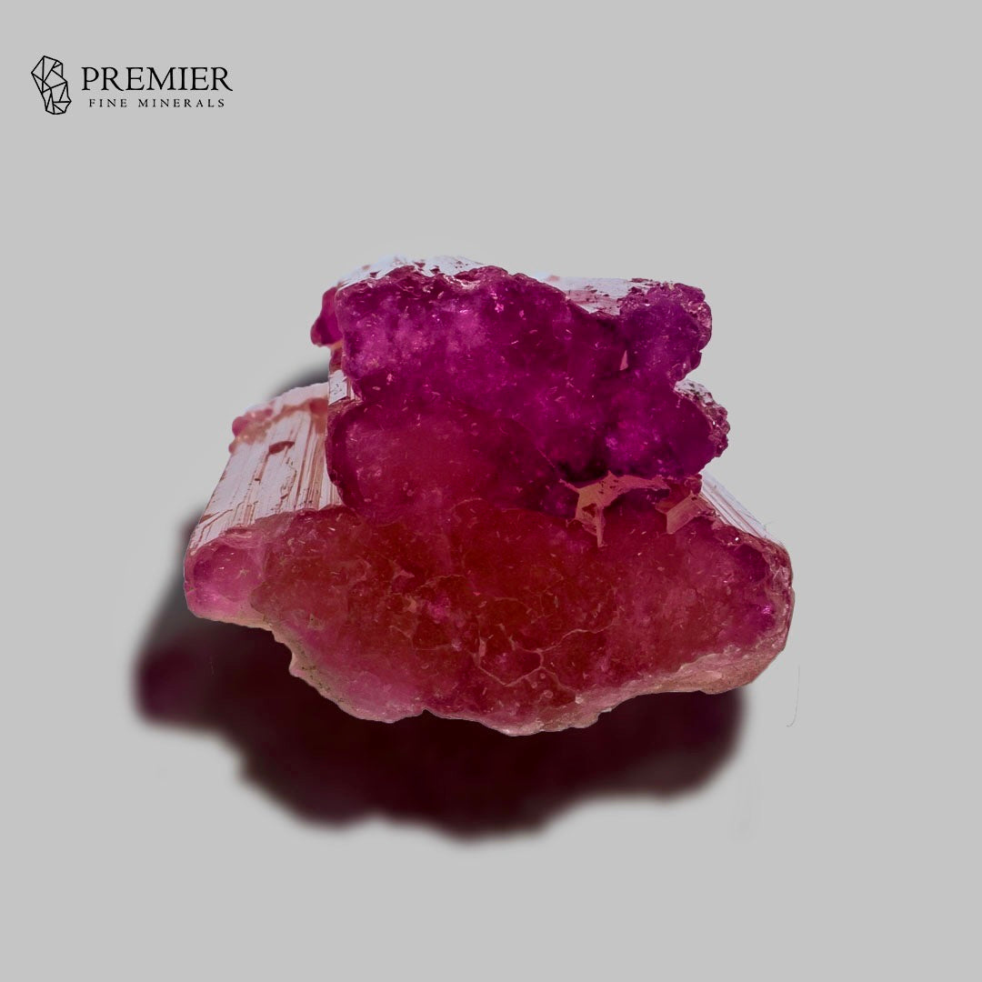 A raw pinkish-red mineral specimen with a bright magenta crystalline top, photographed on a neutral gray background with a shadow. The Premier Fine Minerals logo is visible in the top-left corner.