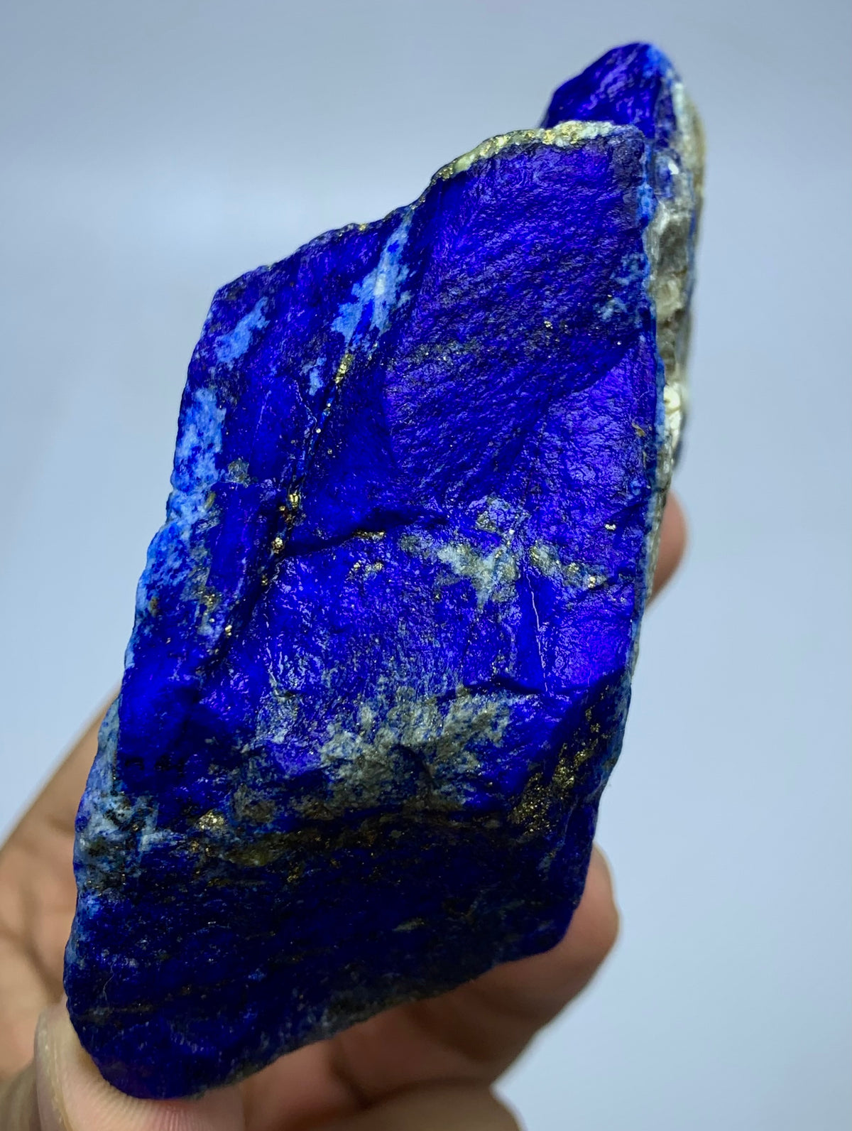 A hand holding a raw, rough blue lapis lazuli crystal with visible gold pyrite veins against a light gray background.