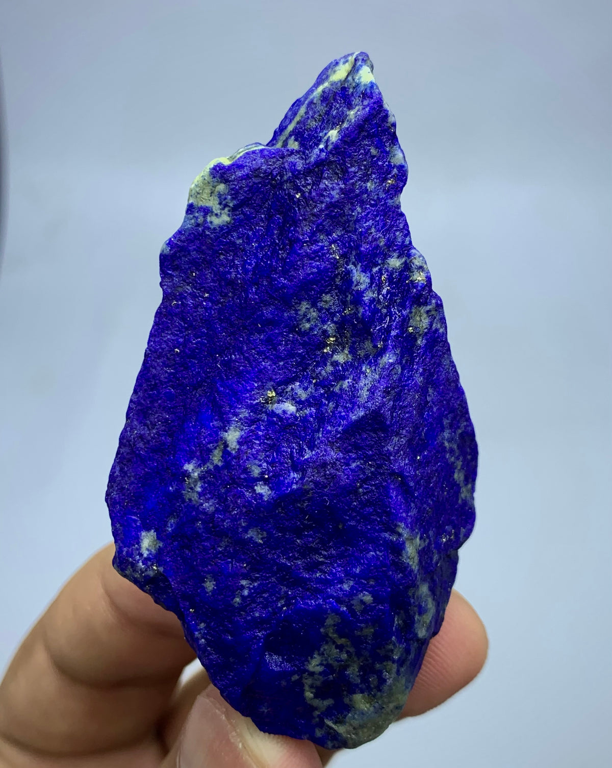 A hand holding a rough, natural lapis lazuli crystal specimen with deep blue coloration and white and pale green veining, shown against a light gray background.