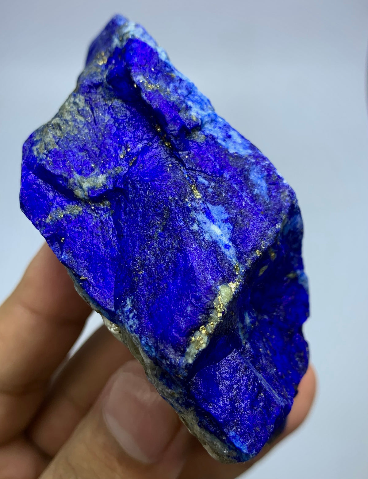 A raw, rough blue lapis lazuli crystal held in a person's hand, showing vibrant deep blue coloration with metallic gold pyrite veins and a silty white matrix.