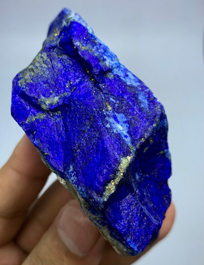 A raw, rough blue lapis lazuli crystal held in a person's hand, showing vibrant deep blue coloration with metallic gold pyrite veins and a silty white matrix.