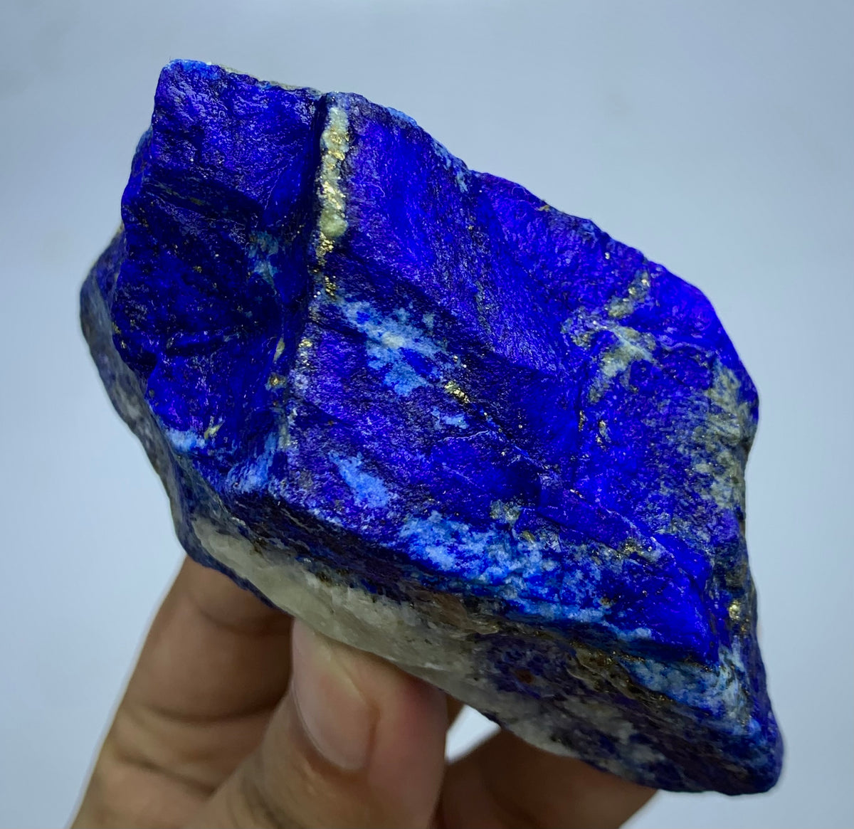 A raw, rough blue lapis lazuli crystal held in a person's hand against a plain light gray background.