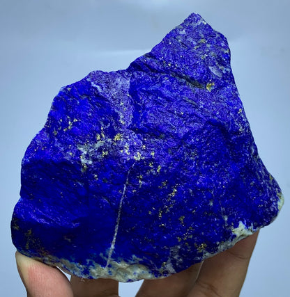 A raw, rough-cut lapis lazuli crystal specimen held up by a hand, showcasing its deep blue color with natural gold pyrite inclusions and a white matrix at the base, set against a plain light gray background.