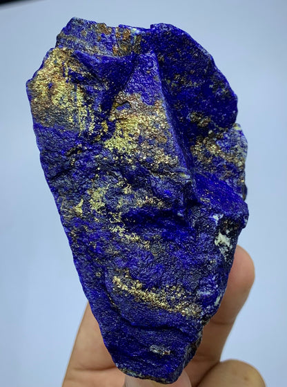 A raw blue lapis lazuli crystal specimen with prominent metallic gold pyrite inclusions, held in a person's hand against a light blue background.