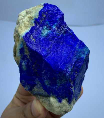 A hand holding a raw blue stone specimen, likely lapis lazuli, showing a vivid deep blue matrix with lighter blue and beige crystalline areas and a rough natural texture.