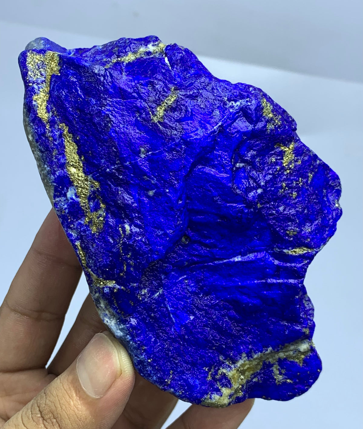 A hand holding a raw, rough-cut lapis lazuli crystal specimen showing deep blue coloration with metallic gold veins, set against a plain light background.