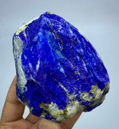  raw lapis lazuli crystal specimen with deep blue, white, and gold matrix, held in a person's hand against a light gray background.