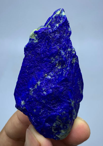A raw, rough-cut lapis lazuli crystal held up by a person's fingers against a light gray background.