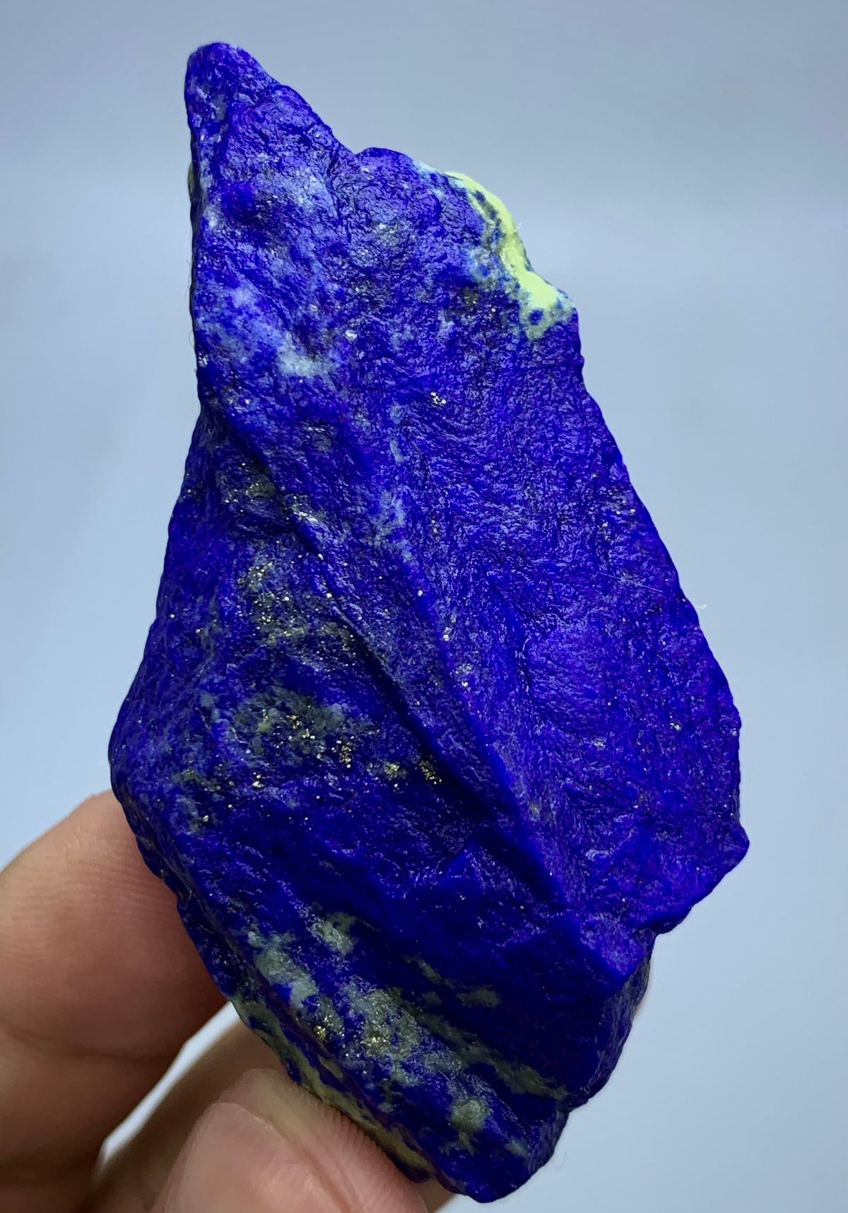 A raw, rough-cut lapis lazuli crystal held in a person's hand, showcasing its deep blue color with natural white and pale yellow matrix textures and metallic flecks.