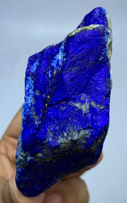 A raw, rough-cut blue lapis lazuli crystal specimen held in a hand against a light gray background.