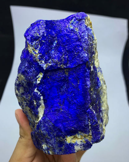 A hand holding a rough, textured lapis lazuli crystal with deep blue coloration and metallic gold pyrite inclusions, set against a white sheet of paper background.