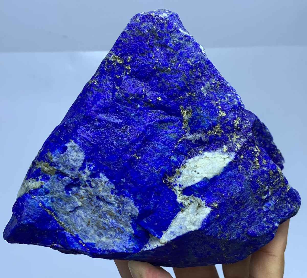 A triangular raw mineral specimen, likely lapis lazuli, held in a hand. The crystal features a vibrant deep blue color with prominent white veining and scattered metallic gold pyrite inclusions.