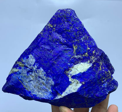 A triangular blue lapis lazuli crystal specimen with gold pyrite veins and white calcite inclusions, held in a hand against a light blue background.