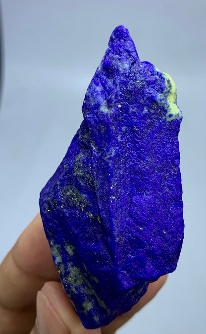 A hand holding a rough, unpolished lapis lazuli crystal with deep blue and purple tones, dark veins, and small golden flecks, against a plain light gray background.